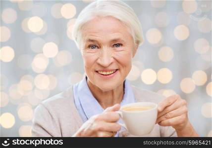 age, drink and people concept - happy smiling senior woman with cup of coffee over holidays lights background