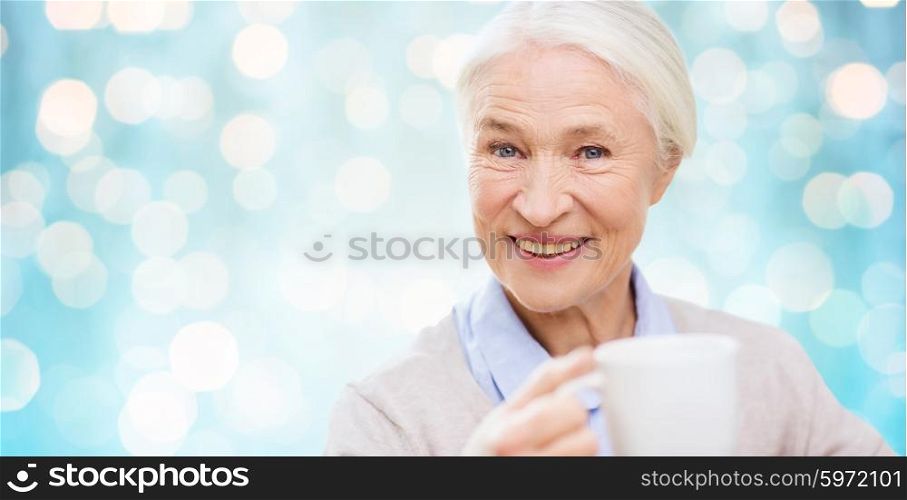 age, drink and people concept - happy smiling senior woman with cup of tea or coffee over blue holidays lights background