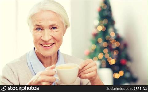 age, drink, holidays, winter and people concept - happy smiling senior woman with cup of coffee at home over living room and christmas tree background