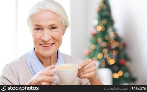 age, drink, holidays, winter and people concept - happy smiling senior woman with cup of coffee at home over living room and christmas tree background