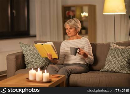 age, leisure and people concept - happy senior woman reading book and drinking coffee or tea at home in evening. senior woman reading book and drinking tea at home