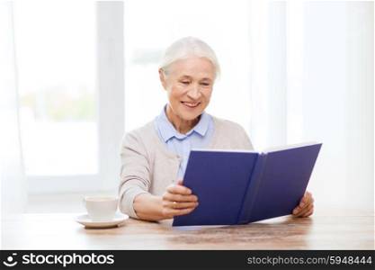 age, leisure and people concept - happy smiling senior woman with cup of coffee reading book at home