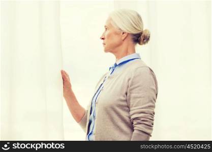 age, loneliness and people concept - lonely senior woman looking through window at home