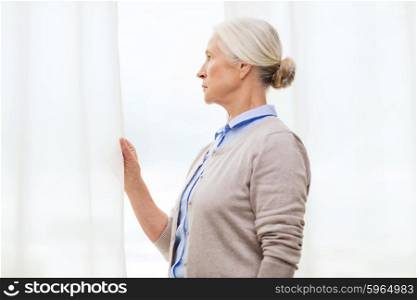 age, loneliness and people concept - lonely senior woman looking through window at home