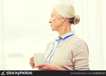 age, loneliness and people concept - lonely senior woman with cup of tea or coffee looking through window at home