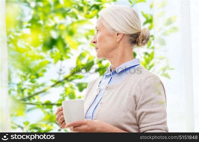 age, loneliness and people concept - lonely senior woman with cup of tea or coffee looking through window at home over window and green natural background. lonely senior woman with cup of tea or coffee