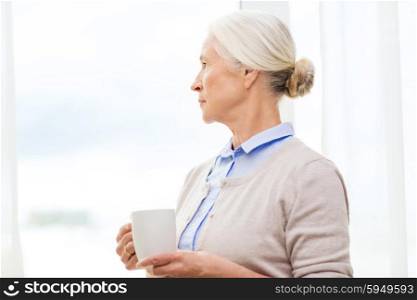 age, loneliness and people concept - lonely senior woman with cup of tea or coffee looking through window at home