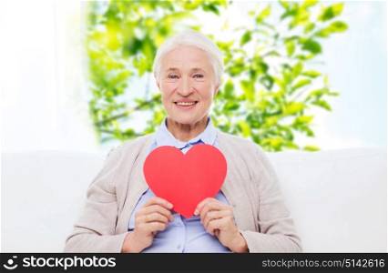 age, love and people concept - happy smiling senior woman with red heart shape at home over window and green natural background. happy smiling senior woman with red heart at home