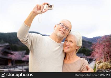 age, tourism, travel, technology and people concept - senior couple with camera taking selfie over asian village landscape background