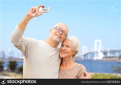 age, tourism, travel, technology and people concept - senior couple with camera taking selfie on city street over rainbow bridge in tokyo and river background