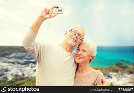 age, tourism, travel, technology and people concept - senior couple with camera taking selfie on street over beach background