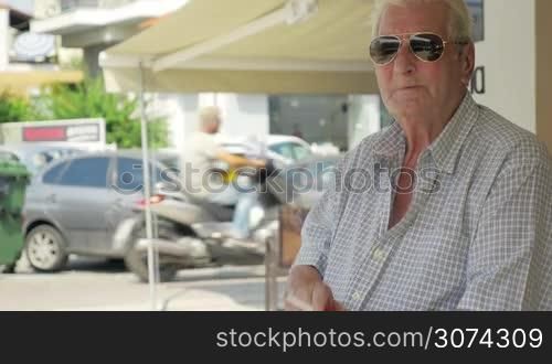 Aged man in sunglasses sitting in the city street and smoking. Enjoying bad habit and observing outside world