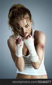 Aggressive female fighter with bruises wearing bloody bandage on her fists, standing in boxing defense position, ready to fight on a neutral grey studio background.