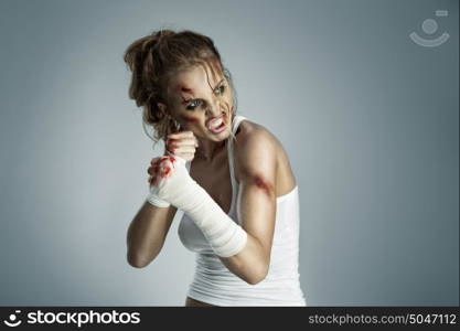 Aggressive female fighter with bruises wearing bloody bandage on her fists, standing in boxing defense position, ready to fight on a neutral grey studio background.