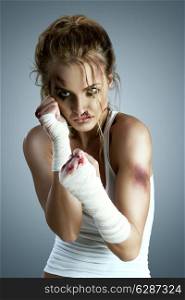 Aggressive female fighter with bruises wearing bloody bandage on her fists, standing in boxing defense position, ready to fight on a neutral grey studio background.