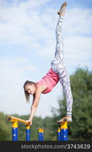 Agile attractive young female gymnast balancing on brightly coloured cross bars outdoors in a park with her leg raised high in the air