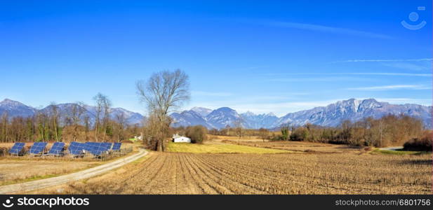 Agricultural winter panorama. Small solar power station. Italian Alps in the background