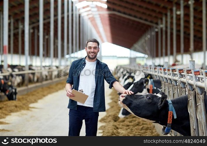 agriculture industry, farming, people and animal husbandry concept - happy smiling young man or farmer with clipboard and cows in cowshed on dairy farm