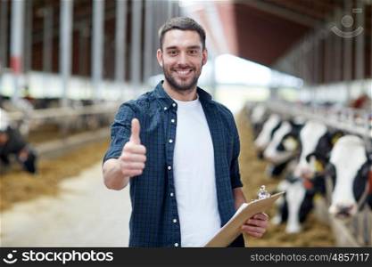 agriculture industry, farming, people and animal husbandry concept - happy smiling young man or farmer with clipboard and cows in cowshed on dairy farm showing thumbs up hand sign
