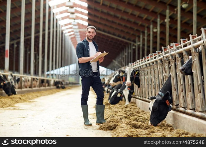 agriculture industry, farming, people and animal husbandry concept - happy smiling young man or farmer with clipboard and cows in cowshed on dairy farm