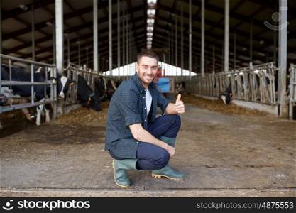 agriculture industry, farming, people and animal husbandry concept - happy smiling young man or farmer with herd of cows in cowshed on dairy farm showing thumbs up hand sign