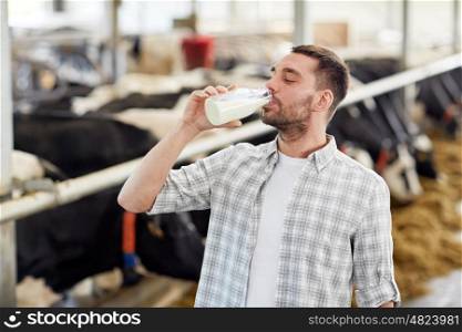 agriculture industry, farming, people and animal husbandry concept - happy young man or farmer drinking cows milk from bottle in cowshed on dairy farm