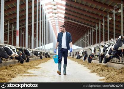 agriculture industry, farming, people and animal husbandry concept - happy young man or farmer with bucket of hay walking along cowshed and cows on dairy farm