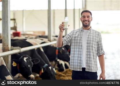 agriculture industry, farming, people and animal husbandry concept - happy young man or farmer with bottle of cows milk in cowshed on dairy farm