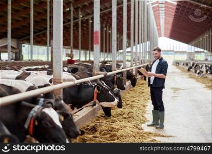 agriculture industry, farming, people and animal husbandry concept - young man or farmer with clipboard and cows eating hay in cowshed on dairy farm