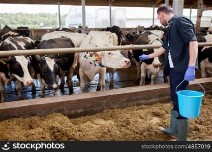 agriculture industry, farming, people and animal husbandry concept - young man or farmer with cows and bucket in cowshed on dairy farm