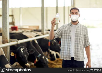 agriculture industry, farming, people and animal husbandry concept - young man or farmer with bottle of cows milk in cowshed on dairy farm wearing medical mask for protection from virus disease. man or farmer in mask with milk on dairy farm