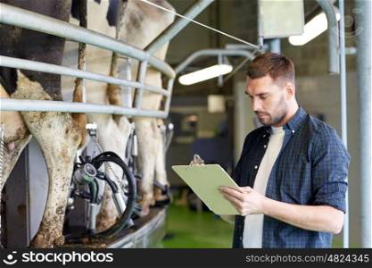 agriculture industry, farming, people, milking and animal husbandry concept - young man or farmer with clipboard and cows at rotary parlour system on dairy farm