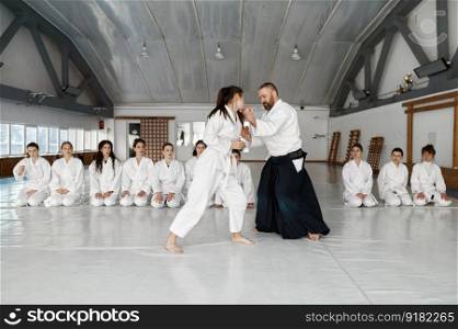 Aikido instructor teaching teenager fighters during group training in gym of sport school. Martial arts master and girl in kimono showing protection technique for students. Aikido instructor teaching teenager fighters during group training in gym