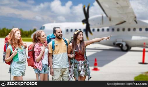 air travel, tourism and adventure concept - group of smiling friends with backpacks pointing finger to something over plane on airfield background. friends with backpacks over plane on airfield. friends with backpacks over plane on airfield