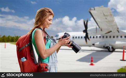air travel, tourism and trip concept - smiling tourist woman with backpack and camera over plane on airfield background. woman with camera and backpack traveling by plane. woman with camera and backpack traveling by plane