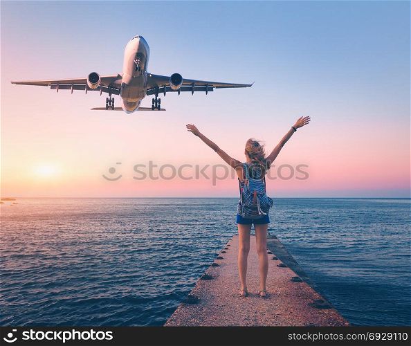 Airplane and happy woman at sunset. Summer landscape with girl standing on the sea pier with raised up arms and flying passenger airplane. Woman and landing commercial aircraft. Lifestyle and travel