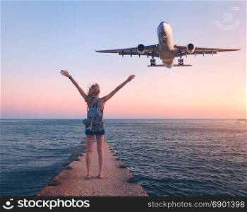 Airplane and woman at sunset. Summer landscape with girl standing on the sea pier with raised up arms and flying passenger airplane. Woman and landing commercial plane in the dusk.Lifestyle and travel