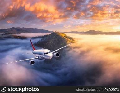 Airplane is flying above mountain peak in low clouds at sunrise. Landscape with passenger airplane, colorful clouds, hills, sky. Aircraft is taking off. Business travel. Commercial plane. Aerial view
