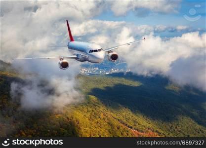 Airplane is flying in clouds over mountains with forest at sunset. Landscape with white passenger airplane, cloudy sky and green trees. Passenger aircraft is landing. Business travel. Commercial plane