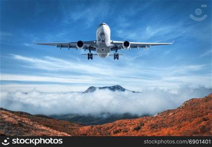 Airplane is flying over low clouds and mountains with autumn forest. Amazing landscape with passenger airplane, trees, mountains, blue cloudy sky. Passenger aircraft. Business travel. Commercial plane
