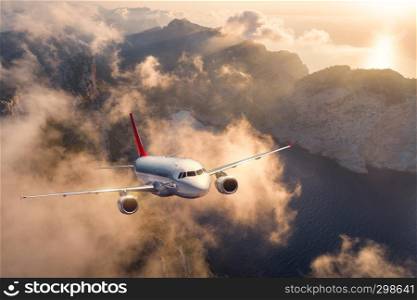 Airplane is flying over mountains and low clouds at sunset in summer. Landscape with passenger airplane, sky in clouds, rocks, sea, sunlight. Business travel. Commercial plane. Aerial view of aircraft
