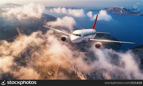Airplane is flying over mountains and low clouds at sunset in summer. Landscape with passenger airplane, sky in clouds, rocks, sea, sunlight. Business travel. Commercial plane. Aerial view of aircraft