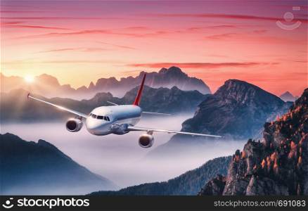 Airplane is flying over mountains in fog at colorful sunset in summer. Landscape with passenger airplane, hills in low clouds, red sky. White aircraft. Business travel. Commercial plane. Aerial view