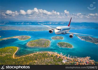 Airplane is flying over small islands and sea at sunny day in summer. Aerial view of passenger airplane, tropical seashore, mountains with green trees, sky and blue water. Top view of aircraft. Travel