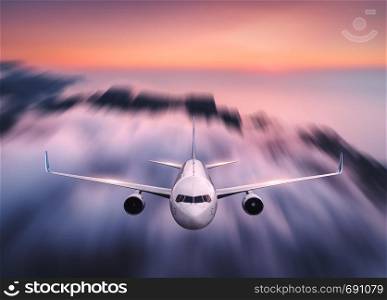 Airplane with motion blur effect is flying over low clouds at sunset. Passenger airplane, blurred clouds, colorful sky at dusk. Aerial view of the modern aircraft. Business travel. Commercial plane
