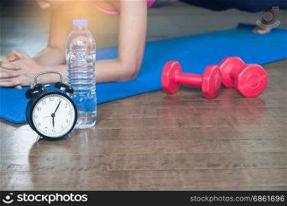 Alarm clock, bottle of water, dumbbells and asian woman yoga at home in background, Working out and lifestyle concept, Selective focus