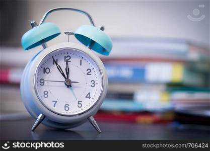 Alarm clock is standing on a grey desk, books and office stuff in the blurry background