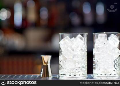 alcohol drinks and luxury concept - two vintage glasses with ice cubes and jigger on bar counter. glasses with ice and jigger on bar counter