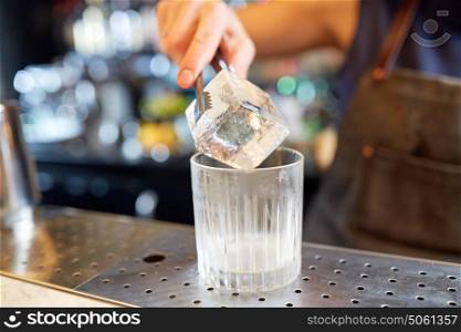 alcohol drinks, people and luxury concept - bartender with tongs adding ice cube into glass and preparing cocktail at bar counter. bartender adding ice cube into glass at bar