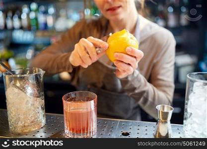 alcohol drinks, people and luxury concept - woman bartender with glass and peeler removing peel from orange and preparing cocktail at bar. bartender peels orange peel for cocktail at bar